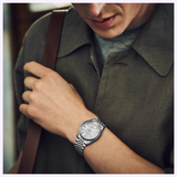 A person wearing a gray collared shirt and carrying a brown strap bag looks down at their left wrist, showcasing the elegance of Japanese craftsmanship with the Seiko Luxe Watches Presage Classic Series HCC001 by Seiko Luxe Watches.