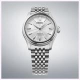 The Seiko Luxe Watches Presage Classic Series HCC001 is an automatic wristwatch featuring a metal link band, minimalist silver hour markers, and a light sunburst dial, beautifully presented against a light gray gradient background—a refined example of Japanese craftsmanship in mechanical watch design.