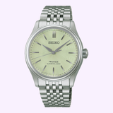 The Seiko Luxe Watches Presage Classic Series HCC002 is a silver automatic wristwatch featuring a pale green textured dial, dark hands, and a metal link bracelet, showcased against a light purple background.