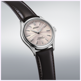 The Seiko Luxe Watches Presage Classic Series HCC003 showcases a Sakura-iro dial with a subtle light pink textured finish, complemented by silver hour markers and a dark brown leather strap, beautifully presented on a gradient light grey background—an exquisite testament to Japanese mechanical watchmaking by Seiko Luxe Watches.