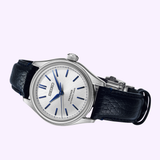 The Seiko Luxe Watches Presage Classic Series HCC004 Limited Edition features a silver textured dial, blue markers and hands, and a black leather strap, shown on a light purple background. This automatic timepiece exemplifies Seiko's elegance and precision.