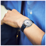 A person in a blue shirt wearing the Seiko Luxe Watches Presage Classic Series HCC004 Limited Edition with automatic movement and a blue leather strap adjusts their bag. The round face displays simple hour markers and dark hands.