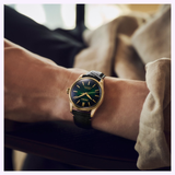 A person wearing a Seiko Luxe Watches Presage Classic Series HCC009 Limited Edition with a green face and black leather strap, resting their arm on a chair, dressed in a beige long-sleeve shirt—a timeless scene that beautifully highlights the elegance of Japanese craftsmanship.