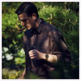 A man wearing a brown button-up shirt and a gold Seiko Luxe Watches Presage Classic Series HCC009 Limited Edition—an elegant example of Seiko Luxe Watches’ Japanese craftsmanship in automatic timepieces—stands outdoors among green trees, looking downward with a thoughtful expression.