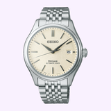 The Seiko Luxe Watches Presage Classic Series SPB463 features an automatic movement, silver metal band, beige dial with a date window at 3 o'clock, and black markers and hands, shown against a light purple background.