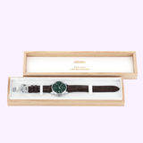The Seiko Luxe Watches Presage Green Enamel Limited Edition SPB111 for men showcases a striking green enamel dial and a brown leather strap, beautifully presented in an open wooden box.