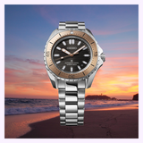 The Seiko Luxe Watches Prospex Sea SPB485, featuring a silver and gold-tone bezel and metal bracelet, is shown against a beach sunset with waves and a vibrant sky.