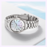 The Seiko Watches Prospex Sea SUR605, a silver watch with a stainless steel band and mother-of-pearl dial, is shown on a white surface against a blurred blue and white background.