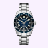 The Seiko Luxe Watches Prospex U.S. Special Edition SPB421 features a stainless steel case, blue bezel, blue textured dial, silver hour markers, date at 3 o’clock, and metal bracelet, shown against a light purple background.