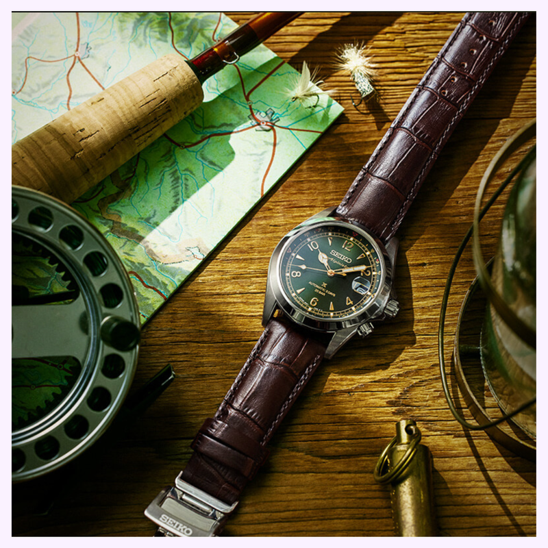 Seiko Watches Prospex Alpinist SPB507 with brown leather strap resting on wood next to a fishing rod, map, and reel, highlighting the adventurous spirit of Seiko Luxe Watches