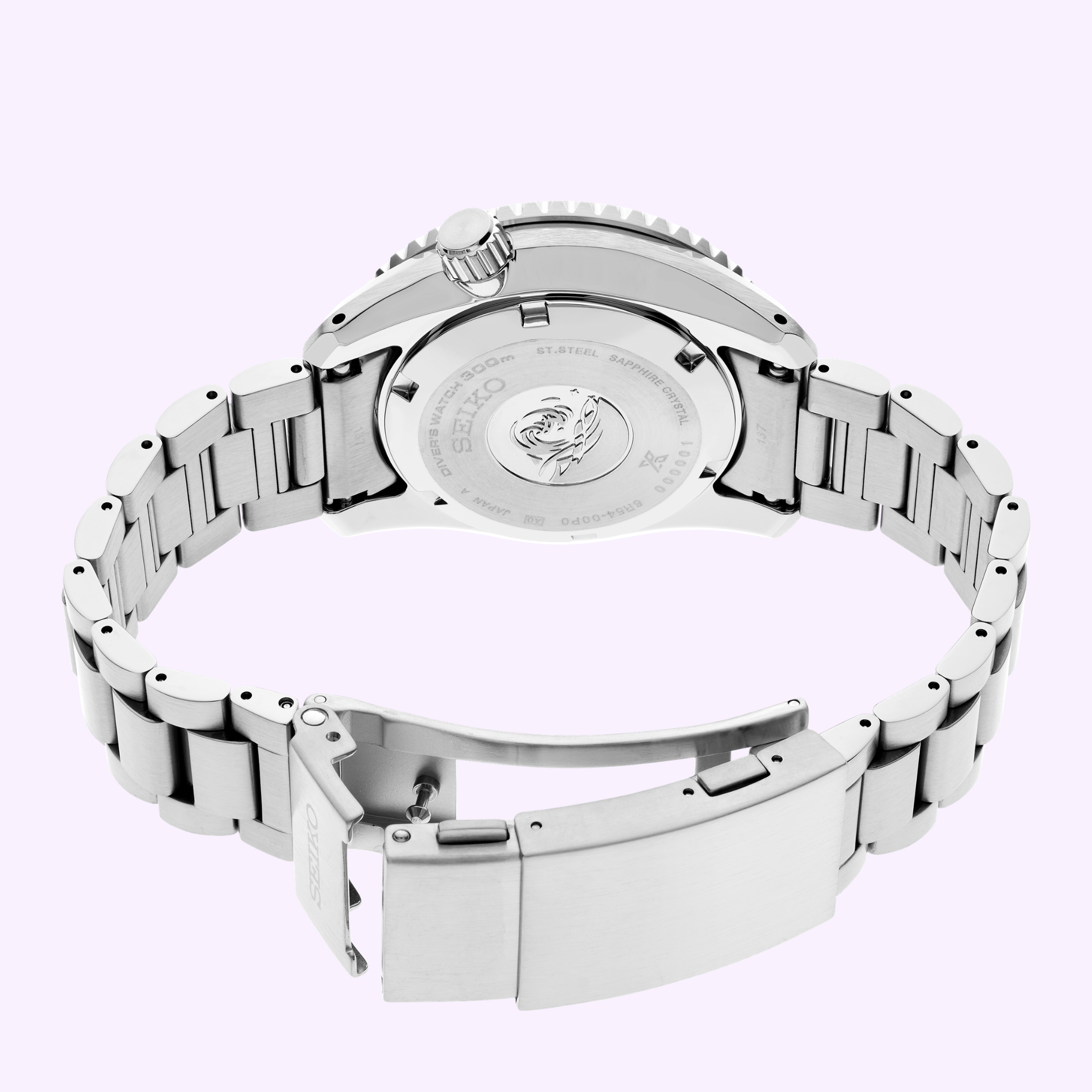 The Seiko Luxe Watches SPB519 PROSPEX HERITAGE DIVER'S GMT, a stainless steel dive watch, is shown with an open clasp revealing the engraved Seiko logo and wave design on the case back, displayed on a light purple background.