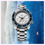 The Seiko Luxe Watches SPB519 PROSPEX HERITAGE DIVER'S GMT sits centered over an aerial beach view, with waves on one side and a small sailboat in the ocean.