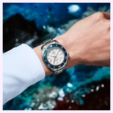 A person in a light blue shirt wears the Seiko Luxe Watches SPB519 PROSPEX HERITAGE DIVER'S GMT—a silver watch with a white dial and blue bezel—standing before a modern, abstract background inspired by diver’s heritage.