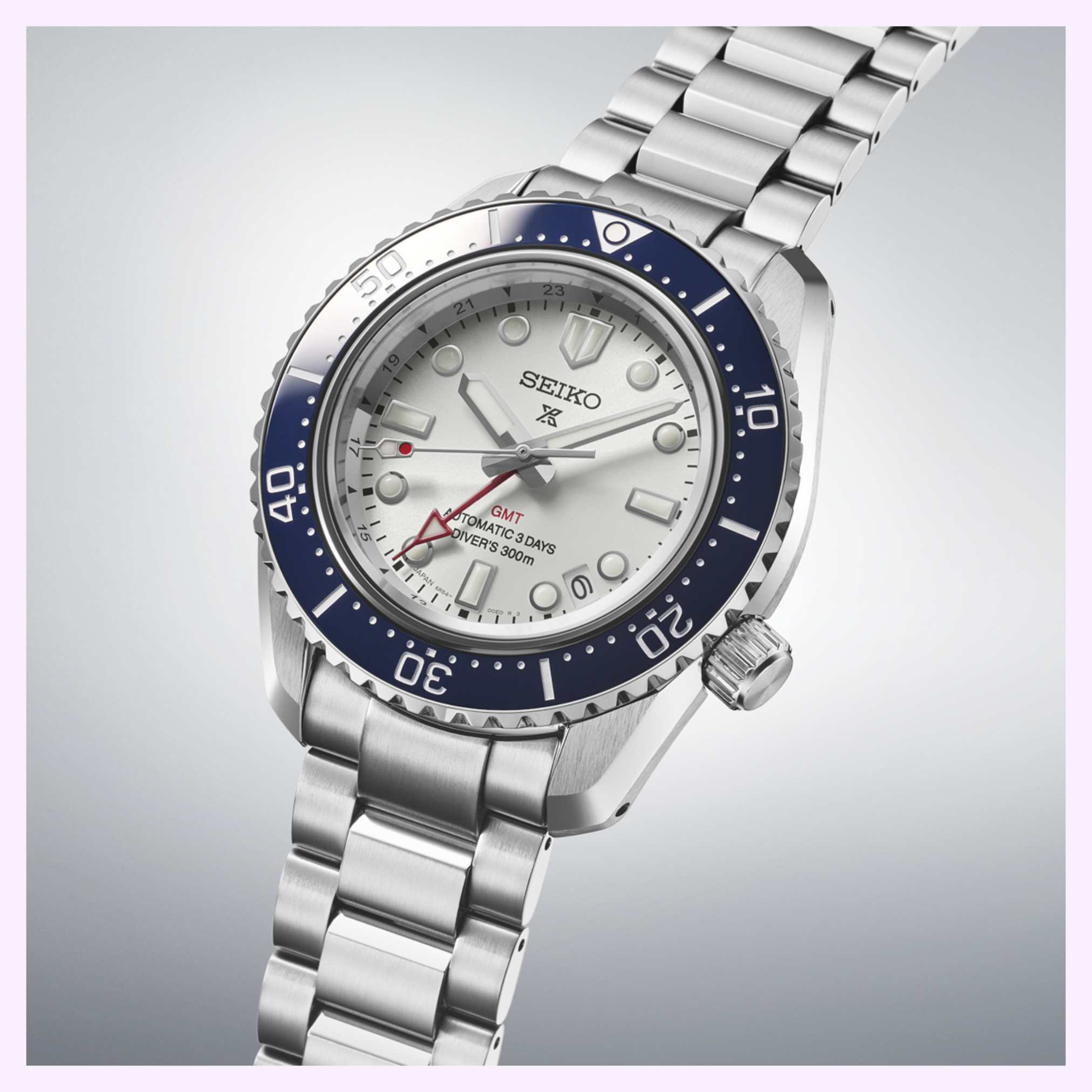 The Seiko Luxe Watches SPB519 Prospex Heritage Diver’s GMT, featuring a silver bracelet, white dial, blue bezel, and red GMT hand, is displayed at an angle on a light gradient background.