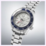 The Seiko Luxe Watches SPB519 Prospex Heritage Diver’s GMT, featuring a silver bracelet, white dial, blue bezel, and red GMT hand, is displayed at an angle on a light gradient background.