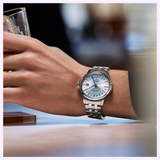 A person wearing a Seiko Luxe Watches SSK037 Presage Cocktail Time with a white dial and Roman numerals holds a glass of whiskey, capturing the timeless elegance of Seiko Luxe Watches against a blurred background.