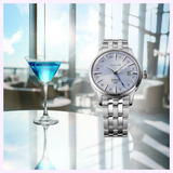 A Seiko Luxe Watches SSK037 Presage Cocktail Time watch with a light blue dial sits next to a blue martini on a reflective table, set against a bright modern lounge with large windows softly blurred in the background.