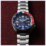 A Seiko Watches 5 Sports SRPD53 automatic watch featuring a blue and red bezel, blue dial, and polished stainless steel band is displayed on a textured red surface. The timepiece displays both the day and date.