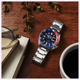 A Seiko Watches 5 Sports SRPD53 by Seiko Watches, featuring a stainless steel case and an eye-catching blue and red bezel, sits on a wooden table next to a notebook, eyeglasses, a sprig of greenery, and a cup of black coffee.