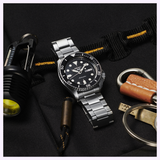 A silver Seiko Watches 5 Sports SSB430 automatic watch with a black dial rests on a black fabric surface, next to a flashlight, a brown leather keychain, and keys.