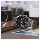 A stainless steel Seiko Watches 5 Sports SSB430 by Seiko Watches, featuring a black dial and bezel, rests on a wooden surface with photos, a camera, and a coffee container in the background.
