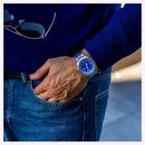 Dressed in a blue sweater and jeans, an individual stands with one hand tucked casually in their pocket, displaying the Seiko Watches 5 Sports X Rowing Blazers SRPM19 Limited Edition—a striking vintage motorsport-inspired timepiece from Seiko Watches featuring a blue dial. In their other hand, they hold a pair of sunglasses, capturing an effortlessly cool aesthetic that pays tribute to the exclusive Seiko 5 Sports collaboration with Rowing Blazers.