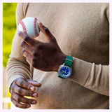 A person wearing a tan sweater and the Seiko Watches 5 Sports X Rowing Blazers SRPM19 Limited Edition vintage motorsport watch by Seiko Watches holds a baseball in one hand while standing outdoors, with blurred greenery in the background.