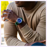 A person in a tan sweater stands outdoors, holding a baseball in one hand while wearing the Seiko Watches 5 Sports X Rowing Blazers SRPM19 Limited Edition on the other wrist—a striking timepiece by Seiko Watches, distinguished by its blue face and green strap, channeling classic vintage motorsport watch style against a bright, blurred background.