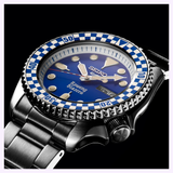 The Seiko Watches 5 Sports X Rowing Blazers SRPM19 Limited Edition wristwatch, crafted by Seiko Watches, showcases a striking blue and white checkered bezel, a vivid blue dial with Rowing Blazers branding, and a silver metal band. The face features both day and date displays. Inspired by the vintage motorsport aesthetic, this limited edition timepiece is presented at an angle against a black background.