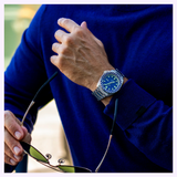 Dressed in a blue sweater, a person adjusts their sleeve with one hand while holding sunglasses in the other, showcasing the Seiko Watches 5 Sports X Rowing Blazers SRPM19 Limited Edition by Seiko Watches.