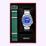 The Seiko Watches 5 Sports X Rowing Blazers SRPM19 Limited Edition is presented in a box, featuring a stainless steel band, a blue dial, and a blue checkered bezel, with an additional green fabric strap placed on the left. The Seiko Watches logo appears at the bottom inside the box.