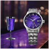 The Seiko Watches Presage Cocktail Time HCB006 145th Anniversary Limited Edition, featuring a striking Edo Purple dial, is displayed beside a purple cocktail in a decorative glass, both set against a softly lit and blurred background.