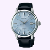 The Seiko Watches Presage Cocktail Time Man SRPB43 by Seiko Luxe Watches is an automatic men's watch with a light blue textured dial, silver hour markers, date window at 3 o’clock, and a black leather strap.