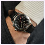 A close-up of a wrist wearing the Seiko Luxe Watches Prospex Alpinist GMT SPB379, featuring a black leather strap and black dial with the time displayed as 10:09. The person is dressed in a light sweater and dark coat.