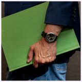 A person in a blue jacket and jeans holds a large green book under their arm, wearing a black Seiko Luxe Watches Prospex Alpinist HBC007 wristwatch with a round face.