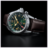 The Seiko Watches Prospex Alpinist HBC007 by Seiko Luxe Watches features a black and green dial with gold hands and indices, a distinctive red GMT hand, reliable Seiko quartz movement, stainless steel case, and a classic brown leather strap, showcased against a sleek black background.
