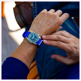 A person wearing the Seiko Watches 5 Sports X Rowing Blazers SRPM21 Limited Edition by Seiko Watches, featuring a round face and blue strap, checks the time while seated with one leg crossed over the other, dressed in blue jeans and a blue sweater.
