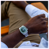 A person wearing the Seiko Watches 5 Sports X Rowing Blazers SRPM21 Limited Edition, featuring a silver case with a green bezel and dark dial, rests their arm on their leg. Dressed in a white shirt and tan pants, they sit on an orange chair, with only part of their body and arm visible to highlight this exclusive Seiko Watches timepiece.
