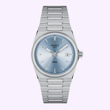 The Tissot Watches PRX 35MM (T1372101135100) features a stainless steel case, light blue dial, silver hour markers, date at 3 o’clock, and a silver metal bracelet, shown on a light purple background.