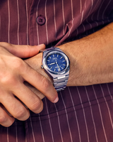 A person wearing a striped maroon shirt displays a Citizen Watches Zenshin Three-hand AW0130-85L silver wristwatch with a blue face and metal band on their left wrist.