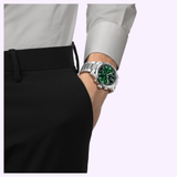 A person dressed in black pants and a light gray shirt stands with their hand in their pocket, showcasing the Tissot Watches Chrono L 42mm T1164171109200 on their wrist. The watch’s green dial catches the eye, while its stainless steel case gleams subtly—highlighting both the elegance and dependable Swiss Quartz movement that Tissot Watches is known for.
