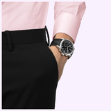 A person in a pink shirt and black trousers has their hand in their pocket, wearing the sleek Tissot Watches Chrono L 42mm T1164171605200 with a round black dial, powered by precise Swiss Quartz movement.