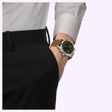 A person wearing black pants and a light gray shirt has their right hand in their pocket, displaying the Tissot Watches Chrono L 42mm T1164171605201, a brown leather wristwatch with a black and yellow face that features the precise Swiss Quartz chronograph movement.