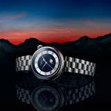 Tissot Watches Desir T1522231149300 watch with a dark blue starry dial, moon phase display, and Roman numeral indices, resting on a reflective surface against a mountain sunset background.