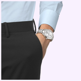A person in a light blue shirt and black pants has their hand in their pocket, showing a Tissot Watches Gentleman 38mm T1658071103100 stainless steel Swiss automatic watch with a white face on their wrist.