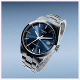 The Tissot Watches Gentleman 38mm T1658071104100 features a silver metallic band, blue dial with silver markers and hands, Powermatic 80 movement, date at 3 o’clock, and “Tissot 1853” on the face, set against a light blue background.
