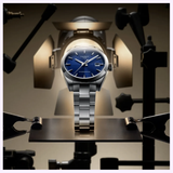 The Tissot Watches Gentleman 38mm T1658071104100, a silver Swiss automatic watch with a blue dial, is displayed on a stand under studio lights, with clamps and equipment visible in the background.
