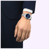 A person in a dark blue suit and white shirt showcases a Tissot Watches Gentleman 38mm T1658071104100 with a blue dial on their left wrist against a light background.