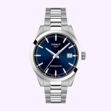 The Tissot Watches Gentleman 38mm T1658071104100 features a silver metal bracelet, blue dial, silver indices and hands, and a date window at 3 o’clock—an elegant Swiss automatic timepiece set against a light purple background.