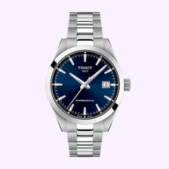 The Tissot Watches Gentleman 38mm T1658071104100 features a silver metal bracelet, blue dial, silver indices and hands, and a date window at 3 o’clock—an elegant Swiss automatic timepiece set against a light purple background.
